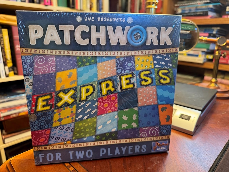 May include: A square board game box titled Patchwork Express for two players by Uwe Rosenberg. The box features a colourful quilt pattern with various fabric designs including fish, flowers, and geometric shapes, with a blue button icon.