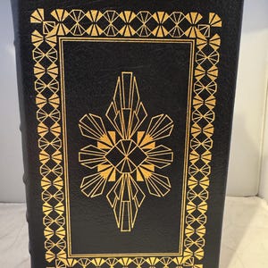 May include: A black leather-bound book with a gold geometric design on the cover. The design features a central diamond shape with radiating lines and a gold border.