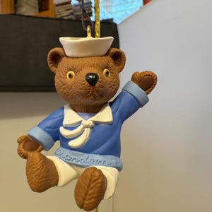 Könnte beinhalten: Ein brauner Teddybär-Ornament, der einen blauen Matrosenanzug und eine weiße Matrosenmütze trägt. Der Bär hat eine weiße Fliege und das Wort "Cherylann" steht unten. Der Bär winkt mit dem rechten Arm.