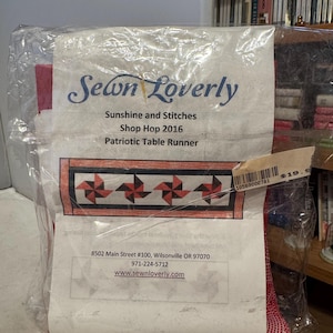 May include: A packaged patriotic table runner featuring a pinwheel design in red, black, and white. The packaging includes the text "Sewn Loverly," "Sunshine and Stitches," and "Shop Hop 2016."