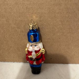 Pode incluir: Enfeite de Natal decorativo em forma de soldado quebra-nozes. O enfeite apresenta um chapéu azul, casaco vermelho, calças pretas e detalhes dourados. O quebra-nozes tem barba branca e uma expressão severa. Uma argola dourada no topo para pendurar.