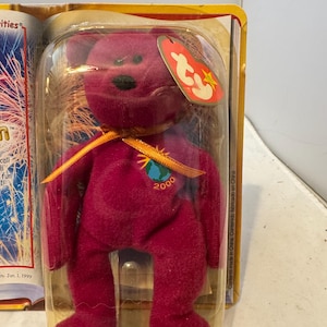 Puede incluir: Un oso de peluche Ty Beanie Baby de color magenta, con una cinta dorada y un parche "2000". El oso está en una funda de plástico transparente con una etiqueta roja y amarilla. El embalaje incluye el texto "Contents made in China."