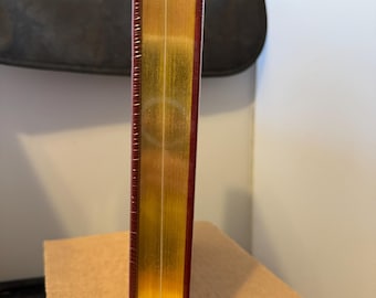 May include: A burgundy hardcover book with gold-gilded edges and a gold spine. The book is wrapped in clear plastic.