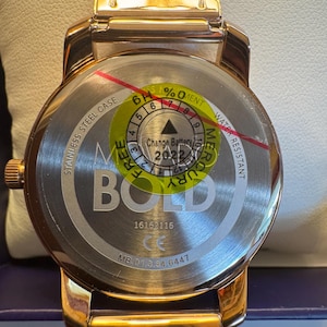 May include: A rose gold watch with a mesh band. The watch face is visible, with the text "BOLD" and "STAINLESS STEEL CASE". The back of the watch has a sticker indicating the battery change year is 2022.