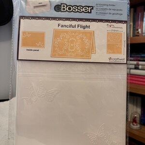 May include: A clear plastic embossing folder package with the text "Bosser" and "Fanciful Flight". The package includes a sample design with butterflies and measurements of 8.27 inches by 11.69 inches. The package also includes the text "craftwell".