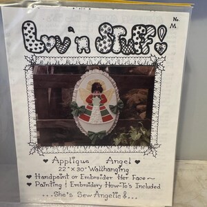 May include: A craft pattern for an applique angel wallhanging. The pattern is 56 cm by 76 cm and features a hand-painted or embroidered angel with a red and green dress. The pattern includes instructions for painting or embroidery.
