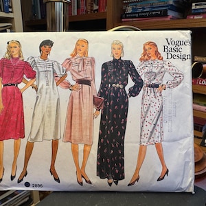 Peut inclure: Une pochette de patron de couture en papier blanc intitulée Vogue's Basic Design 2896, présentant des illustrations de cinq styles de robes différents avec diverses longueurs de manches, encolures et détails de taille, incluant des motifs unis et floraux.