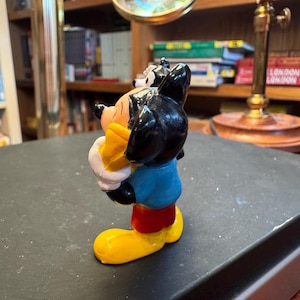 Puede incluir: Una colorida figura de Mickey Mouse. La figura tiene orejas negras, una cara amarilla y viste una camisa azul, pantalones cortos rojos y zapatos amarillos. La figura sostiene un objeto amarillo.