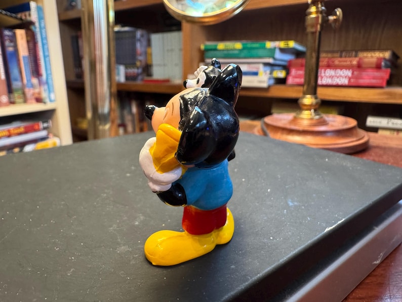 Puede incluir: Una colorida figura de Mickey Mouse. La figura tiene orejas negras, una cara amarilla y viste una camisa azul, pantalones cortos rojos y zapatos amarillos. La figura sostiene un objeto amarillo.