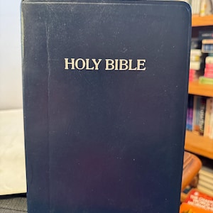 Könnte beinhalten: Eine marineblaue Bibel mit dem weißen Aufdruck "HOLY BIBLE". Das Buch weist eine leicht abgenutzte Erscheinung auf. Im Hintergrund ein Bücherregal.