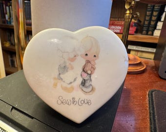 Puede incluir: Caja de baratijas de cerámica en forma de corazón con una ilustración pintada de dos niños y un oso de peluche. La caja es blanca con un tinte rosado y las palabras "Sew in Love" están escritas en la parte delantera.