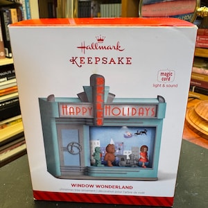 May include: A Hallmark Keepsake Window Wonderland Christmas tree ornament box featuring a teal storefront with a marquee reading Happy Holidays and Belles. The display window shows a robot, teddy bear, train, and doll with a Santa sleigh silhouette.