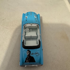 May include: A blue toy car with a white roof and a black interior. The car features a picture of Elvis Presley on the back with his signature. The car has a guitar design on the bonnet.