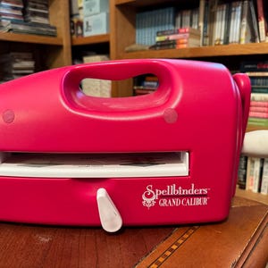 Może przedstawiać: Jasnoróżowa maszyna do wycinania i wytłaczania Spellbinders Grand Calibur. Maszyna ma białą rączkę i biały wałek. Maszyna stoi na drewnianej powierzchni.