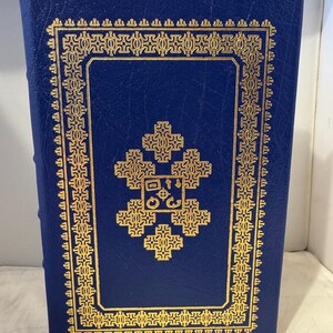 Può includere: Un libro rilegato in pelle blu con un design dorato sulla copertina. Il design presenta una croce stilizzata con un cerchio al centro. Il libro è circondato da un bordo dorato.