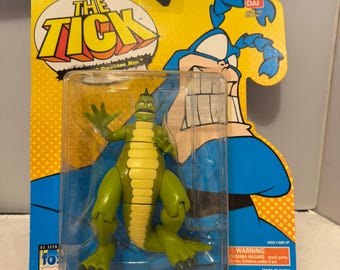 May include: A green and yellow Dinosaur Neil action figure from The Tick, packaged on a yellow and blue backing card. The figure is in a clear plastic case. The packaging includes the text "The Tick" and "Dinosaur Neil".