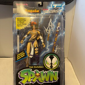 Könnte beinhalten: Deluxe Ultra-Action-Figur von Angela aus Todd McFarlanes Spawn-Comic-Serie. Die Figur befindet sich in einer versiegelten Verpackung und enthält ein abnehmbares Schwert und einen Stab. Die Verpackung zeigt das Spawn-Logo und Text.