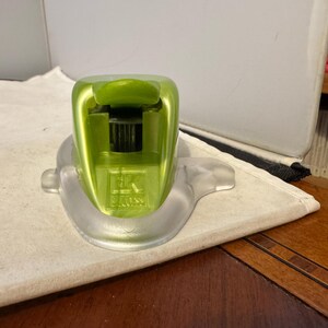 May include: A lime green stapler with a clear plastic base. The stapler has a rounded top with a lever and a black interior. The base has a curved design. The stapler is sitting on a white surface.