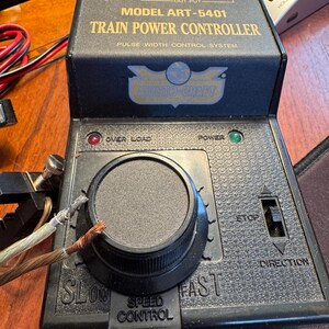 May include: A black Aristo-Craft Model ART-5401 train power controller with a pulse width control system. The device features a large central speed control dial, a direction switch, and indicator lights for power and overload status.