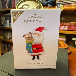 May include: A Hallmark Keepsake Christmas tree ornament box featuring Santa Claus holding a dog in a stocking. The box is white with red and gold accents. The ornament is a 2012 exclusive VIP gift.