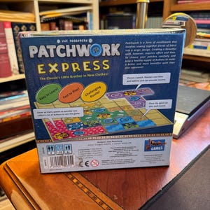 May include: A square board game box titled Patchwork Express by Uwe Rosenberg. The blue box features game instructions, colourful quilt tile illustrations, and text including Easy to Learn, Fast to Play, and Challenging Puzzle. It shows player counts, age ratings, and a barcode on the bottom edge.