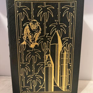 May include: A black leather-bound book with a gold-embossed design featuring a monkey, palm trees, and a rocket ship.