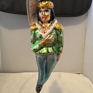 Puede incluir: Adorno navideño de cristal que representa una figura del ballet El Cascanueces. El adorno presenta una chaqueta verde con detalles dorados, pantalones azules y una corona dorada. La cara de la figura está pintada con rasgos delicados.