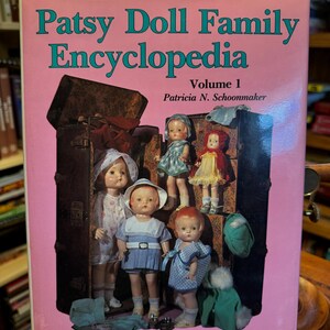 Peut inclure: Couverture de livre rose avec le titre "Patsy Doll Family Encyclopedia Volume 1" en lettres turquoise. La couverture présente des poupées vintage dans différentes tenues, disposées près de valises ouvertes. Le nom de l'auteur, Patricia N. Schoonmaker, est également visible.