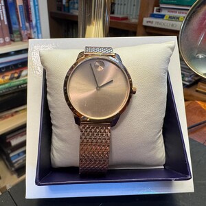 May include: A rose gold-toned wristwatch with a mesh band, displayed on a white pillow inside a blue-trimmed box. The watch has a minimalist design with a circular face and two hands.