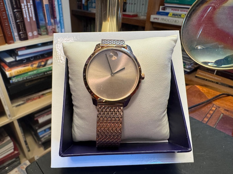 May include: A rose gold-toned wristwatch with a mesh band, displayed on a white pillow inside a blue-trimmed box. The watch has a minimalist design with a circular face and two hands.