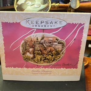 May include: A Keepsake Ornament box featuring a pink and white design with a central oval illustration. The illustration depicts two rabbits in a garden setting, with the text "Garden Bunnies" and "Nature's Sketchbook".