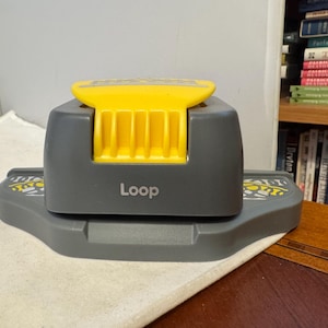 Pode incluir: Uma ferramenta de artesanato cinza e amarela com a palavra "Loop" impressa na lateral. A ferramenta tem uma alça amarela e uma base cinza com padrões decorativos. É projetada para artesanato e projetos de papel.