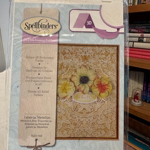 Puede incluir: Un paquete de carpetas de relieve 3D Spellbinders. El paquete presenta un diseño floral con las palabras "We love YOU". El paquete también incluye el texto "Labels 34 Medallion" y "E3D-016".