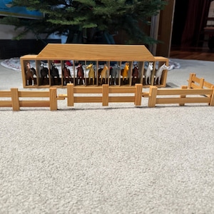 Puede incluir: Un establo de juguete de madera con una fila de caballos en miniatura de varios colores. Una valla de madera está frente al establo. El establo tiene un techo y paredes de color marrón claro. Los caballos están en boxes individuales.
