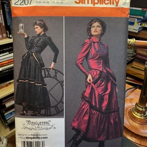 Könnte beinhalten: Simplicity Schnittmuster 2207 mit zwei Frauen in Kleidern im viktorianischen Stil. Das Muster enthält die Größen 14, 16, 18 und 20. Die Kleider sind schwarz und bordeauxrot mit Rüschenröcken und taillierten Jacken. Die Marke des Musters ist Simplicity.