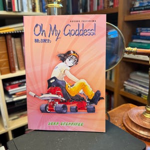 May include: A manga volume titled Oh My Goddess! Sora Unchained by Kosuke Fujishima. The cover features a character with short dark hair wearing a yellow shirt and pants, sitting in a red go-kart against a pink background with radial lines.