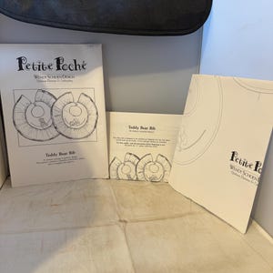 May include: Two white paper patterns for a teddy bear bib. The patterns are from Petite Poche by Wendy Schoen Design. The patterns include instructions and diagrams for sewing the bib.
