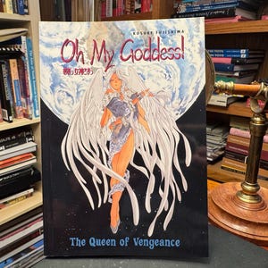 May include: A manga book titled Oh My Goddess! The Queen of Vengeance by Kosuke Fujishima. The cover features a central illustration of a winged figure with long white hair against a large blue and white planet background and a dark space sky.