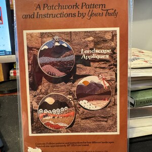 Könnte beinhalten: Ein Vintage-Musterpaket zum Herstellen von vier Landschaftsapplikationen. Das Paket hat einen braunen Hintergrund mit einem Bild von vier fertigen Applikationen, die jeweils eine andere Landschaftsszene darstellen. Das Paket trägt den Titel "Landschaftsapplikationen" und enthält Schnittmuster in Originalgröße und Anleitungen. Die fertige Größe jeder Applikation beträgt etwa 25,4 cm im Durchmesser.