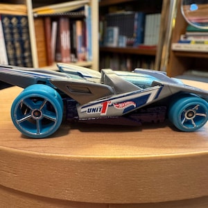 May include: A silver and dark blue toy race car with bright blue wheels. The side of the vehicle features the text UNIT 1 and a red and white Hot Wheels logo. The car has a low-profile, aerodynamic design with a pointed rear section and an open cockpit.