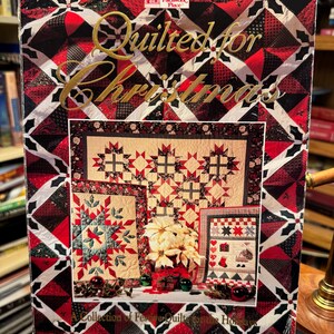Könnte beinhalten: Ein Buch mit dem Titel "Quilted for Christmas" mit einem geometrischen Muster in Rot, Schwarz und Weiß. Das Cover zeigt Bilder von Quilts zum Thema Weihnachten, darunter Stern- und Herzmotive. Das Buch ist eine Sammlung festlicher Quilts für die Feiertage.