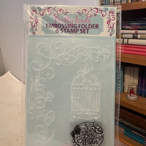 Peut inclure: Un ensemble de gaufrage et de tampons pour l'artisanat dans un emballage plastique transparent. L'ensemble comprend une cage à oiseaux et un motif floral. Le tampon indique "Best Wishes". L'emballage est étiqueté "Pretty Birds EMBOSSING FOLDER & STAMP SET".