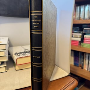 May include: A hardcover book titled "The Compleatest Angling Booke" by Crawhall. The book has a black spine with gold lettering and detailing. The book is in a decorative slipcase with a textured, light-colored design. The publisher is Freshet Press.
