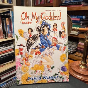 May include: A manga volume titled Oh My Goddess! Ninja Master by Kosuke Fujishima. The cover features a central female character with long dark hair, surrounded by smaller figures and various colorful flowers against a white background.