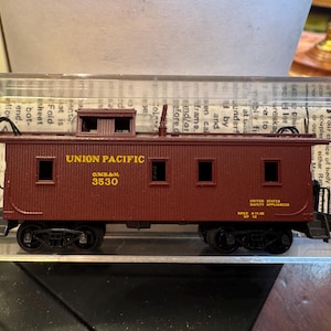 May include: A model train caboose in a deep red color, featuring the text "UNION PACIFIC" and the number "3530" in yellow. The miniature train car has black wheels and small windows, with details like a roof cupola and ladders.