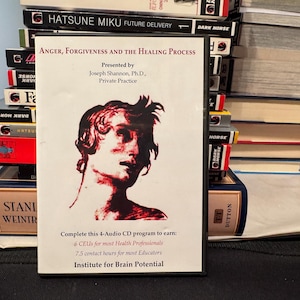 May include: A CD case titled "Anger, Forgiveness and the Healing Process" with a red and white image of a person's face. The case is surrounded by stacks of books. The text on the case says "Presented by Joseph Shannon, Ph.D., Private Practice."