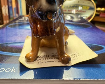 May include: A ceramic Saint Bernard dog figurine with brown and white markings. The dog is sitting on a small paper tag with text. The figurine is on top of a book. The text on the tag reads "AGEN-REN MADE IN SAN DIN".