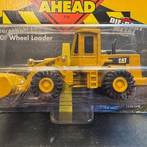 May include: A yellow die-cast metal Caterpillar 950F wheel loader toy in its original clear plastic blister packaging. The packaging features the text AHEAD, Caterpillar 950 Wheel Loader, and CAT. The toy has black tyres and a front bucket.