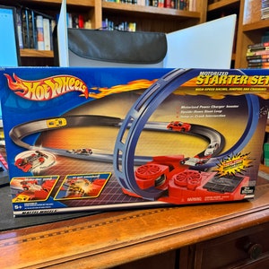Puede incluir: Un set de inicio motorizado Hot Wheels con una pista azul y un impulsor rojo. El set incluye un coche y cuenta con un bucle, un salto y una intersección de choque. La caja dice "Carreras de alta velocidad, saltos y choques".