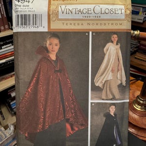May include: Simplicity Vintage Closet sewing pattern featuring cape designs. The pattern includes a red cape with a detailed pattern, a cream-colored cape with fur trim, and a black cape with purple lining. The pattern is labeled "One Size."